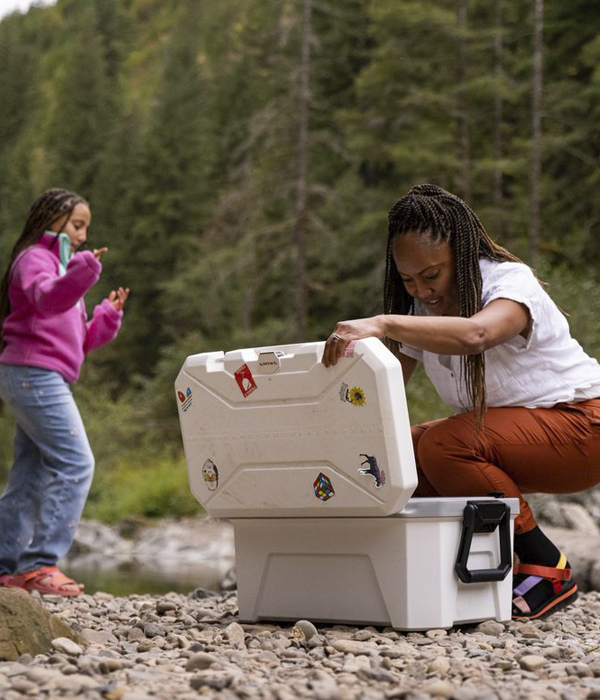 A young black woman peers into an open Plano cooler while her daughter plays behind her on the stony river bank.