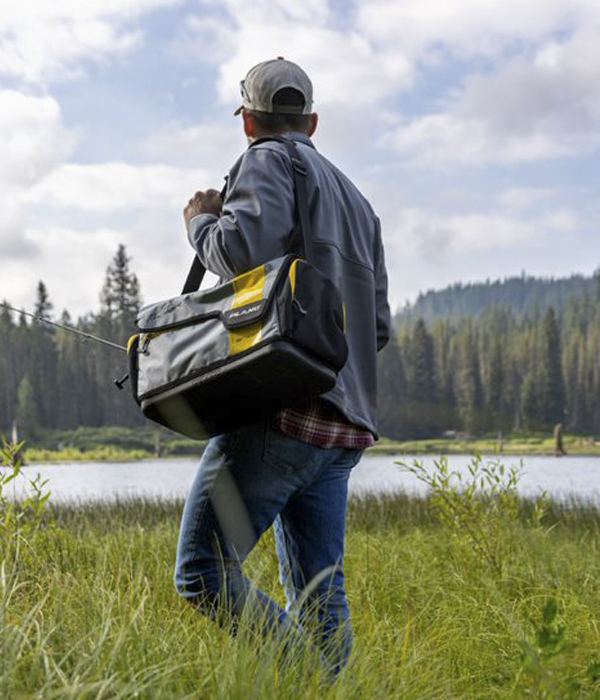 A man wearing flannel and a baseball cap walks through the tall grass growing on the riverside on a cloudy day. There is a soft sided tackle bag slung over his shoulder.