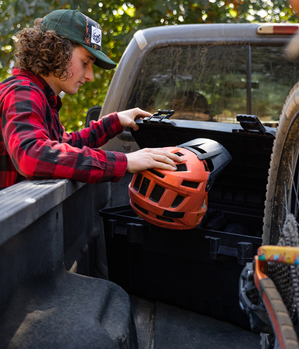 A curly-headed youth in a baseball cap is leaning over the side of a pickup truck bed and is stowing a cycling helmet in a black storage trunk.