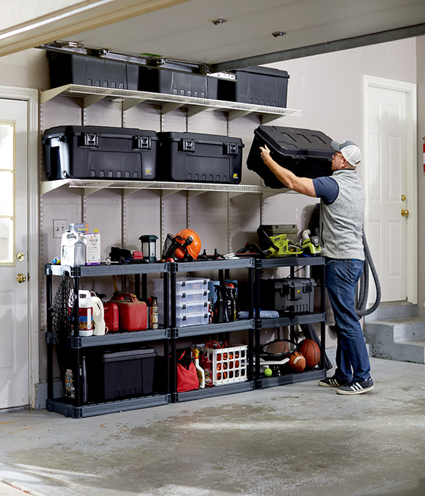 A man is lifting a black storage trunk and storing it on a shelf in his garage.