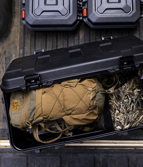 A Plano trunk sits with it's lid ajar on the tailgate of a pickup truck, exposing gear bags and camouflage hunting apparel inside.